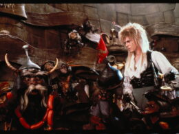 David Bowie as Jareth the Goblin King standing among various goblin puppets on the set of the film Labyrinth.