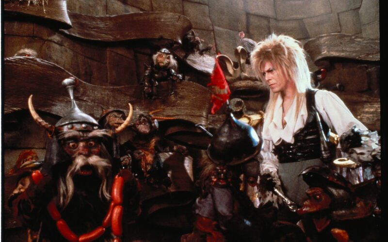 David Bowie as Jareth the Goblin King standing among various goblin puppets on the set of the film Labyrinth.